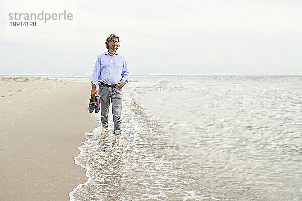 Älterer Mann hält Schuhe in der Hand und läuft an der Küste am Strand entlang