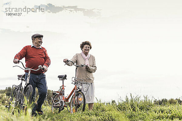 Happy senior couple walking with bicycles under sky