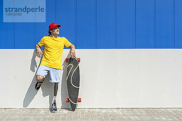 Lächelnder junger Mann mit Behinderung  der an einem sonnigen Tag neben dem Skateboard steht