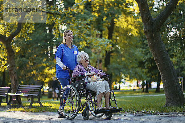 Lächelnde Mitarbeiterin im Gesundheitswesen geht mit einer älteren Frau im Rollstuhl im Park spazieren