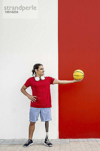 Lächelnder Mann mit Beinprothese hält gelben Basketball an der Wand