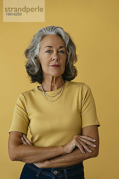 Senior woman standing with arms crossed against yellow background