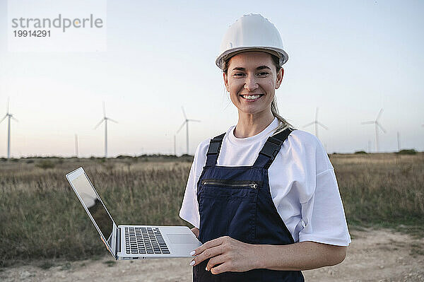 Glücklicher Ingenieur mit Schutzhelm und Laptop vor Windkraftanlagen bei Sonnenuntergang