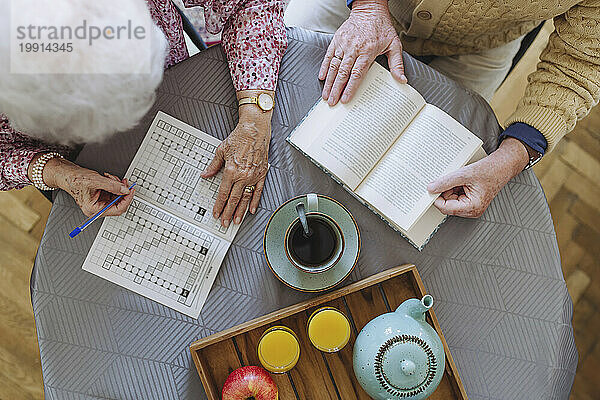 Senior man reading book with woman doing crossword puzzle at table