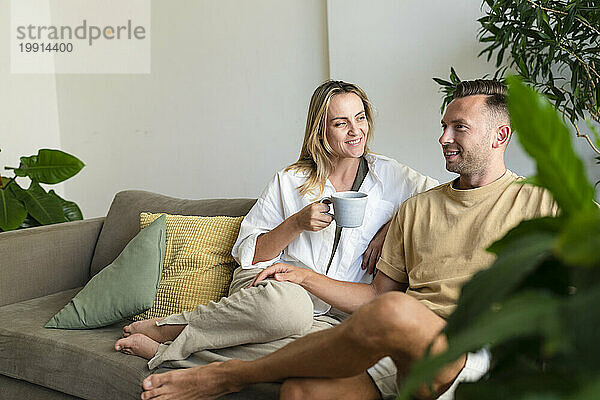 Lächelnde blonde Frau mit Kaffeetasse im Gespräch mit Mann auf dem heimischen Sofa