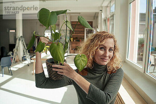 Lächelnde Geschäftsfrau  die Muskeln spielen lässt und eine Topfpflanze im Büro hält