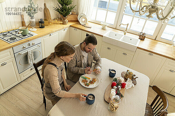 Couple decorating cookies with hot chocolate mugs on table in kitchen