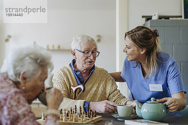 Glücklicher Mitarbeiter im Gesundheitswesen  der einem älteren Paar am Tisch Tee serviert
