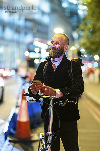 Lächelnder Mann hält Smartphone in der Hand und steht mit Fahrrad in der Stadt