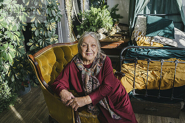 Lächelnde ältere Frau sitzt zu Hause auf einem Sessel neben dem Bett