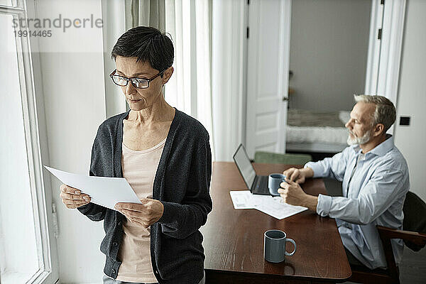 Pensive woman looking at business paper standing next to husband sitting at table working on laptop