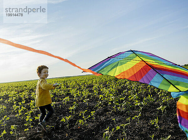 Lächelnder Junge  der Drachen auf einem landwirtschaftlichen Feld unter Himmel steigen lässt