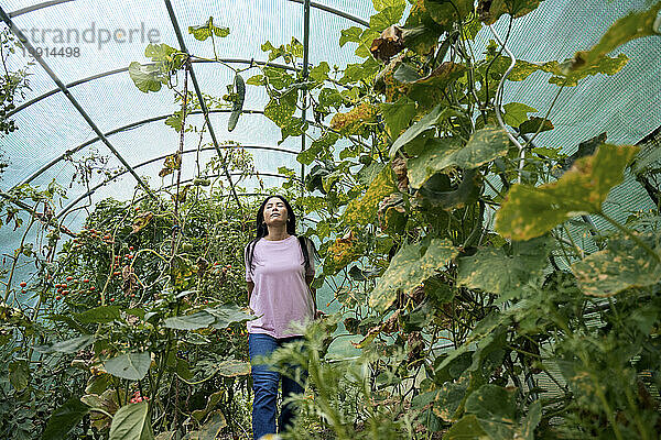 Woman with eyes closed walking amidst plants in greenhouse