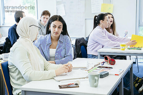 Frau diskutiert mit Freundin im Hijab  die im Klassenzimmer am Schreibtisch sitzt