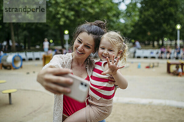 Kleines Kleinkindmädchen und Mutter haben Spaß auf dem Spielplatz und machen Selfies mit dem Smartphone