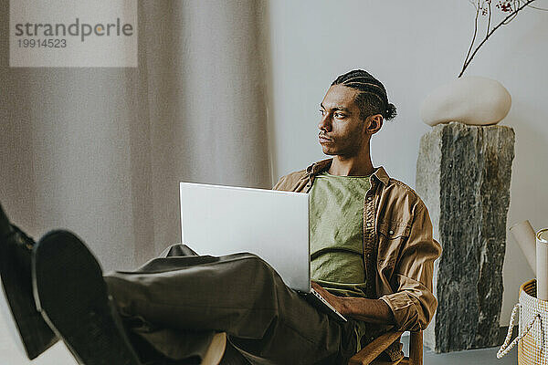 Nachdenklicher junger Mann mit Laptop entspannt sich zu Hause mit Füßen auf dem Tisch