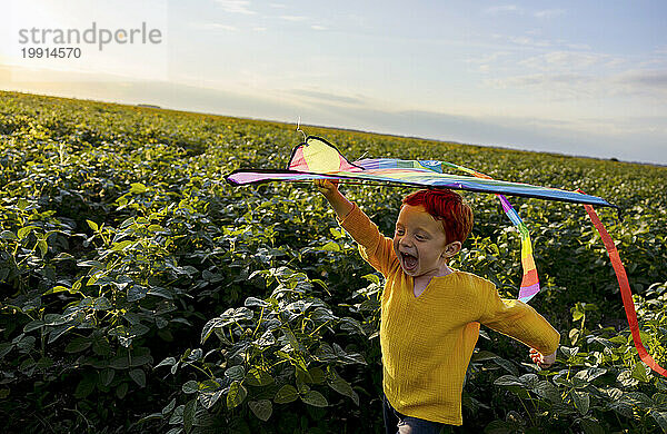Glücklicher Junge hält Drachen auf dem Kopf und rennt auf landwirtschaftlichem Feld