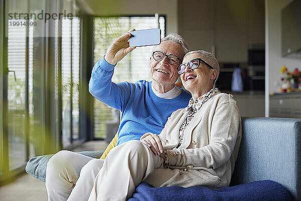 Happy senior couple sitting on couch at home taking selfie