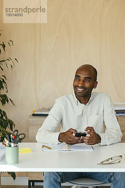 Lächelnder Geschäftsmann sitzt mit Smartphone am Schreibtisch im Heimbüro