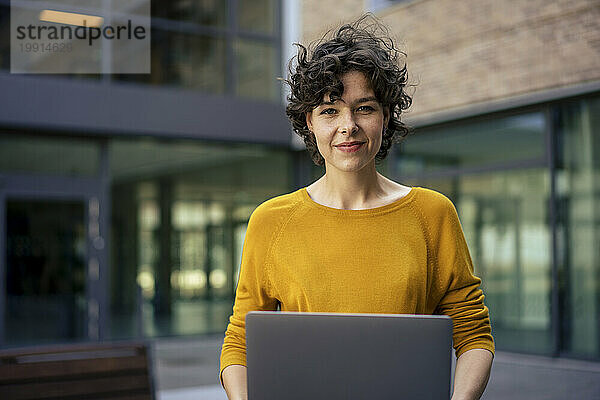 Lächelnde Geschäftsfrau mit Laptop vor Bürogebäude