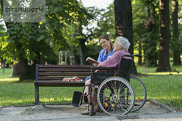 Glückliche Mitarbeiterin im Gesundheitswesen mit einer älteren Frau  die im Rollstuhl im Park sitzt