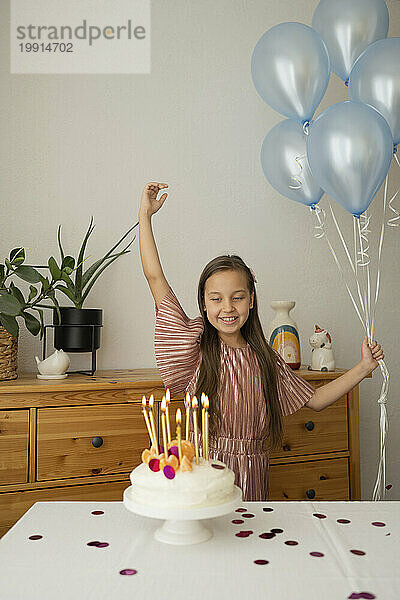 Fröhliches Mädchen hält Luftballons in der Hand und feiert zu Hause Geburtstag mit Kuchen