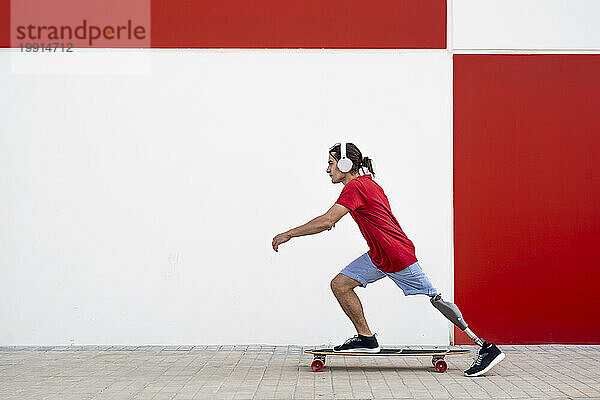 Mann trägt kabellose Kopfhörer und fährt mit einer Beinprothese Skateboard vor der Wand