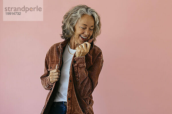 Lächelnde ältere Frau trägt braune Jacke und steht vor rosa Hintergrund