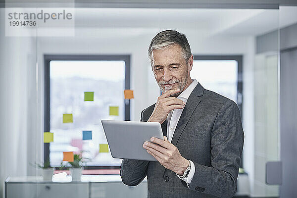 Smiling senior businessman with hand on chin using tablet PC in office