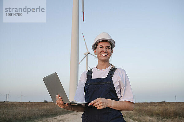 Glücklicher Ingenieur mit Schutzhelm und Laptop vor Windkraftanlagen