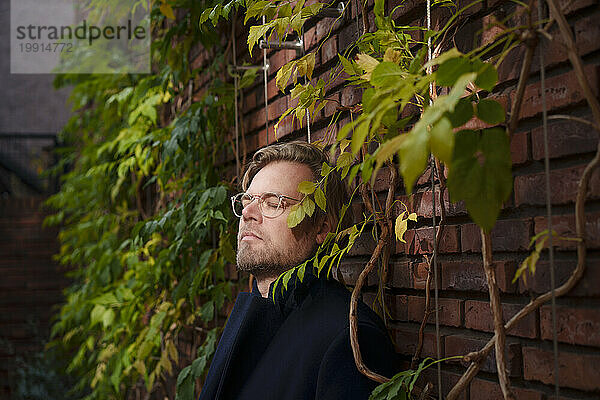 Businessman standing with eyes closed leaning on brick walli overgrown with plants
