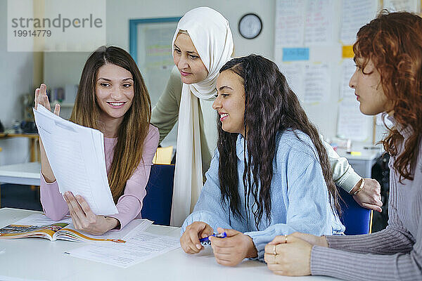 Lächelnde multiethnische Schüler diskutieren über Papier am Schreibtisch im Klassenzimmer