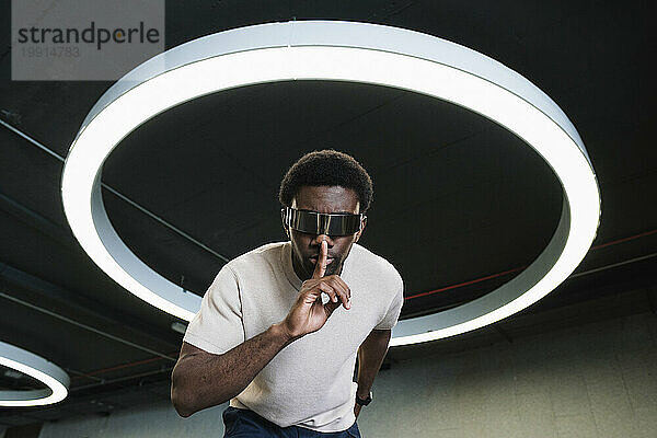 Young man with futuristic cyber glasses under modern ring lamp putting forefinger on lips