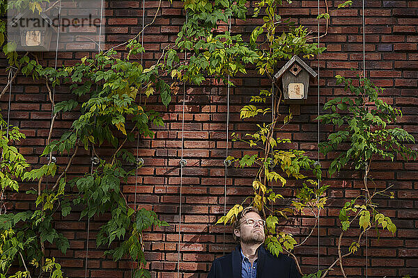 Businessman with eyes closed leaning on brick wall overgrown with plants
