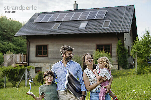 Junge Familie steht vor ihrem Einfamilienhaus mit Sonnenkollektoren auf dem Dach