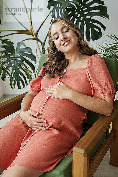 Smiling pregnant woman sitting with eyes closed on armchair at home