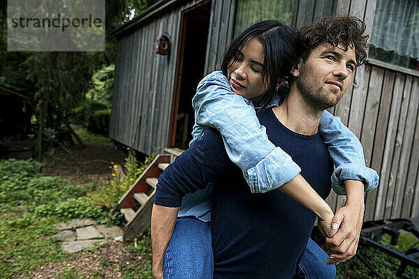 Smiling man giving piggyback ride to woman in front of wooden cabin