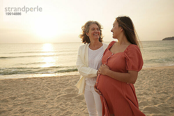 Glückliche Mutter und schwangere Tochter gehen am Strand spazieren