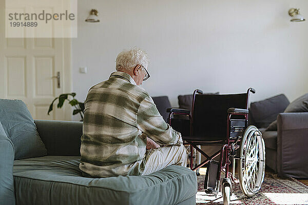 Trauriger älterer Mann sitzt zu Hause auf dem Sofa neben dem Rollstuhl