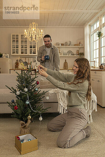 Lächelnde Frau schmückt den Weihnachtsbaum  während ein Mann zu Hause steht