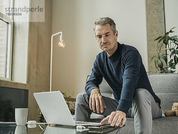 Freiberufler sitzt mit Smartphone und Laptop am Couchtisch