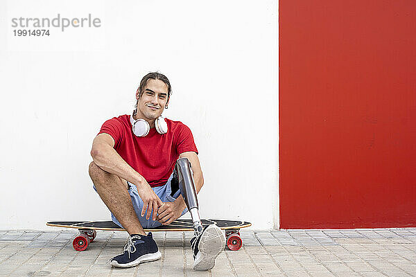 Lächelnder junger Mann sitzt auf Skateboard mit Beinprothese vor der Wand