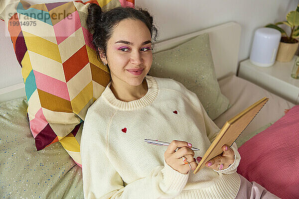 Lächelnde  nachdenkliche Frau mit Buch und Stift  die zu Hause im Bett liegt
