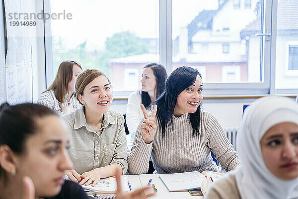 Lächelnder Student gestikuliert Friedenszeichen  während er neben einem Freund im Klassenzimmer sitzt