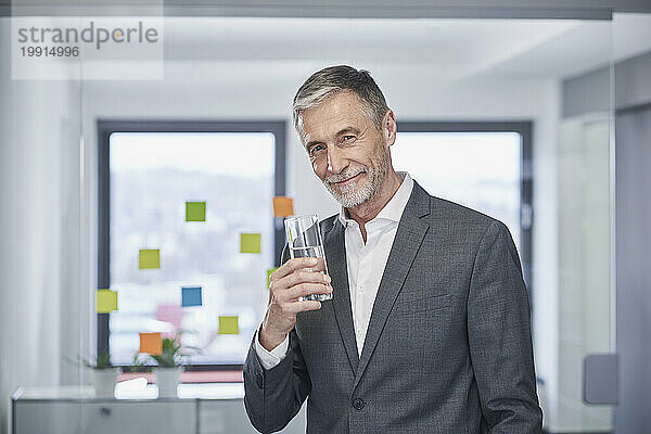 Lächelnder Geschäftsmann steht mit einem Glas Wasser im Büro