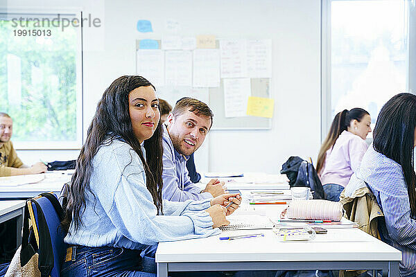 Lächelnde  vielfältige Freunde sitzen am Schreibtisch mit Büchern im Klassenzimmer