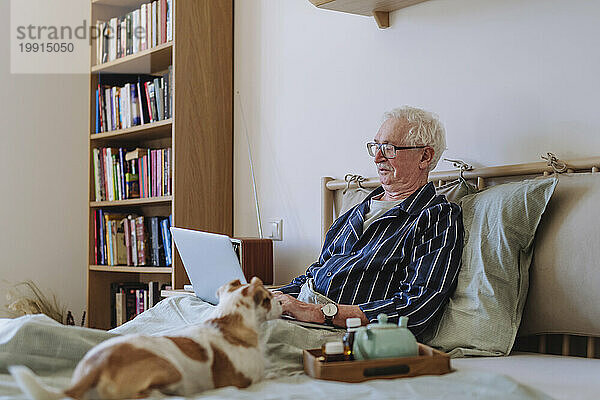 Älterer Mann benutzt Laptop neben Hund im heimischen Bett