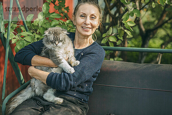 Lächelnde Frau sitzt mit Katze auf Schaukel im Garten