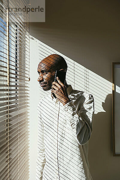 Mann schaut durch Jalousien hinaus und spricht zu Hause mit dem Smartphone