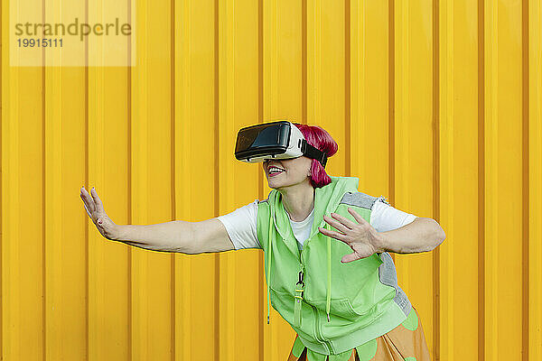 Ältere Frau trägt Virtual-Reality-Simulator und gestikuliert vor gelber Wand